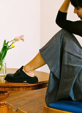 Lade das Bild in den Galerie-Viewer, Frau mit Rock, die in einer Altbauwohnung auf einem Holzstuhl sitzt und ihr linkes Bein locker auf einem Tisch aus Holz abgestellt hat. Auf dem Tisch stehen Tulpen. Sie blickt verträumt, an ihren Füßen sind coole schwarze Clogs aus Leder von Cedoublé zu sehen.