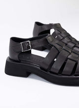 Load image into Gallery viewer, Detailaufnahme von schwarzen Fishermen Sandals aus schwarzem, super weichem Echtleder. Durch die soften Riemen spürt man diesen Schuh kaum am Fuß. Es handelt sich um eine Modellvariante mit geschlossener Hinterkappe und Vorderkappe. Dadurch eignet sich der Schuh auch für Personen, die einen luftig-sommerlichen Schuh wünschen, aber ihre Füße gerne kaschieren. Die Lederriemen sind verflochten, sodass ein korbartiges Dessin entsteht.