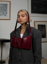 Lade das Bild in den Galerie-Viewer, Eine weinrote Crossbody-Bag aus weich gepolstertem Glattleder mit drei voneinander abgetrennten Fächern hängt um den Kopf eines weiblichen Models mit kinnlangen, blonden Haaren. Das Model trägt einen grauen Oversize-Blazer mit Schulterpolstern.