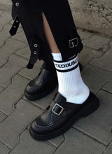 Lade das Bild in den Galerie-Viewer, Bild einer coolen Kombination von weißen Tennissocken mit Logo-Print und hochwertigen schwarzen Leder-Clogs mit weißen Nähten. Die schwarze Hose, die das Model dazu trägt, verfügt über einen Beinschlitz. Dieser klafft offen, sodass der Logo-Schriftzug auf der Socke gut zur Geltung kommt. Sowohl Socke als auch Schuhe sind von der Marke Cedoublé.  Die Clogs verfügen über eine Monk-Schnalle und stehen optische großen Designerlabels in nichts nach.