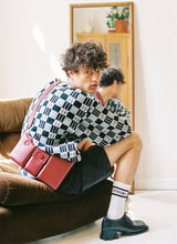 Lade das Bild in den Galerie-Viewer, Cooles Foto eines männlichen Models mit lockigen Haaren und modischem Styling auf einem braunen Cordsofa sitzend. Im Fokus stehen die rote Umhängetasche aus feinem Kalbsleder sowie die schwarzen Lederschuhe mit einem ausgefallenen Absatz. Beide Artikel sind vom deutschen Hersteller Cedoublé.
