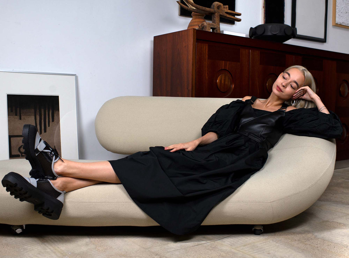 A young woman with chin-long, blonde hair is casually laying on a sculptural-like, off-white couch in a vintage design furniture studio. She is wearing a long, black dress with puffy sleeves and a black harness while looking at her greyish mesh sneakers.