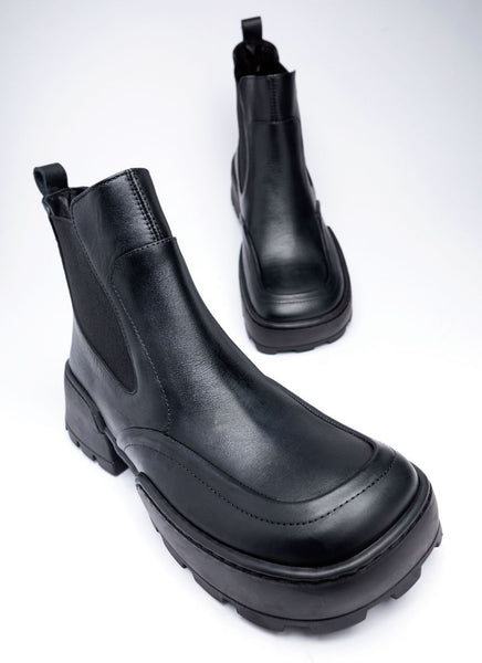 Ein Paar schwarze Boots aus Leder in Karreeform. Die Sohle ist eine dicke Chunky Sohle aus rutschfestem Gummi. In der Mitte ist die Laufsohle dünner, sodass die Abrollbewegung des Fußes erleichtert wird. Die Schuhe verfügen über einen elastischen Elastikeinsatz wie er typisch ist für Chelsea-Boots. 