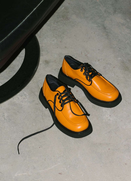 Ein Paar orangefarbener unisex Lederschnürer steht auf einem Betonboden und wird von einem harten Licht angestrahlt