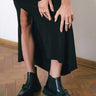 Ein Foto von grazilen Modelbeinen, die in schwarzen Leder Chelsea Boots mit Schleife stecken. Das Model ist sehr edel mit einem langen, schwarzen Kleid mit Schlitz gekleidet. Die Frau zieht mit gepflegten Händen, die von silbernen Ringen geziert werden, das Kleid leicht nach oben, sodass der Fokus auf die hochwertigen Schuhe gelenkt wird. Das Model befindet sich in einem Altbau, es ist ein schönes Fischgrät-Parkett zu sehen.