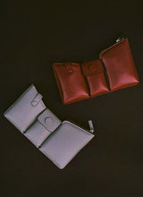Load image into Gallery viewer, Two bags on a black background, one is made out of puffy, dark red leather, the other is made out of grey pebbled leather. The bags have three compartments.
