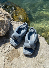 Lade das Bild in den Galerie-Viewer, Sommerliche Sandale aus grauem, weich gepolstertem Leder steht an einem Seeufer in Italien. Diese verschlungenen Knoten Sandals sind von dem jungen, deutschen Schuhlabel Cedoublé.
