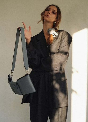 Bild eines weiblichen Models, welches in einen oversized, grauen Herrenanzug gekleidet ist. Ihre langen, braunen Haare sind mit einem grauen Cord-Scrunchie zu einem seitlichen, lockeren Zopf gebunden, sie trägt Silberschmuck. Den linken Arm hält sie vor der Brust verschränkt, der rechte Arm ist angewinkelt. An diesem baumelt locker eine hellgraue, hochwertige Designertasche. Diese ist schmal in der Tiefe und verfügt über Karabiner, sodass der Gurt ausgetauscht werden kann.