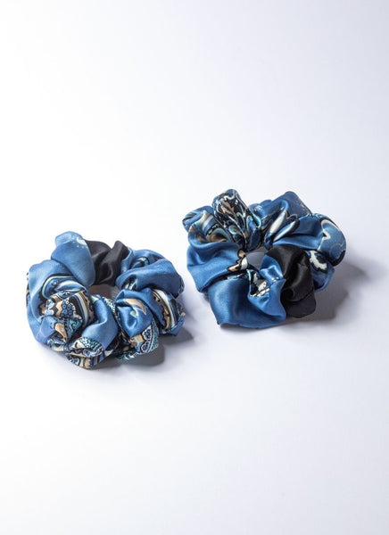 Auf dem Foto sind zwei Scrunchies aus reiner, glänzender Seide in verschieden Blautönen abgebildet. Die Grundfarbe ist ein frisches Himmelblau. Das Muster in beige-weiß-schwarz ist ein klassisches Paisley-Dessin. 